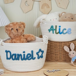 May include: Two white rope baskets with blue embroidered names, "Daniel" and "Alice", each holding a stuffed animal. A wooden bunny rattle and a wooden plaque with a name are also visible. The baskets are on a wooden surface.
