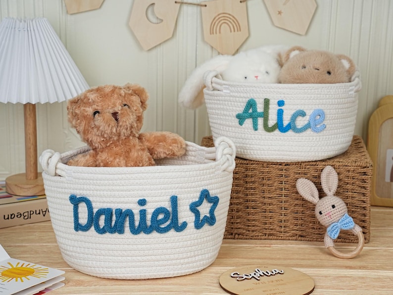 May include: Two white rope baskets with blue embroidered names, "Daniel" and "Alice", each holding a stuffed animal. A wooden bunny rattle and a wooden plaque with a name are also visible. The baskets are on a wooden surface.