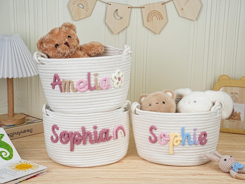 May include: Three white rope baskets with embroidered names: "Amelia", "Sophia", and "Sophie". Each basket contains a stuffed animal. The "Amelia" basket has a flower accent, while the "Sophia" basket has a rainbow accent. The baskets are on a wooden surface.