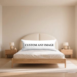 May include: A white body pillow with the text "CUSTOM ANY IMAGE" in black lettering is displayed on a wooden bed frame. The bed is in a neutral-toned bedroom with matching bedside tables and lamps.