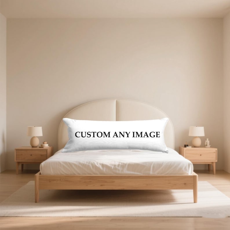 May include: A white body pillow with the text "CUSTOM ANY IMAGE" in black lettering is displayed on a wooden bed frame. The bed is in a neutral-toned bedroom with matching bedside tables and lamps.
