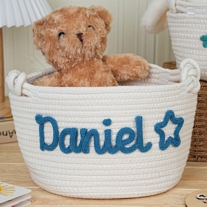 May include: A white rope basket with the name "Daniel" in blue script and a star. A brown teddy bear sits inside the basket. Another basket with the name "Ali" is visible in the background. The baskets are for storage.