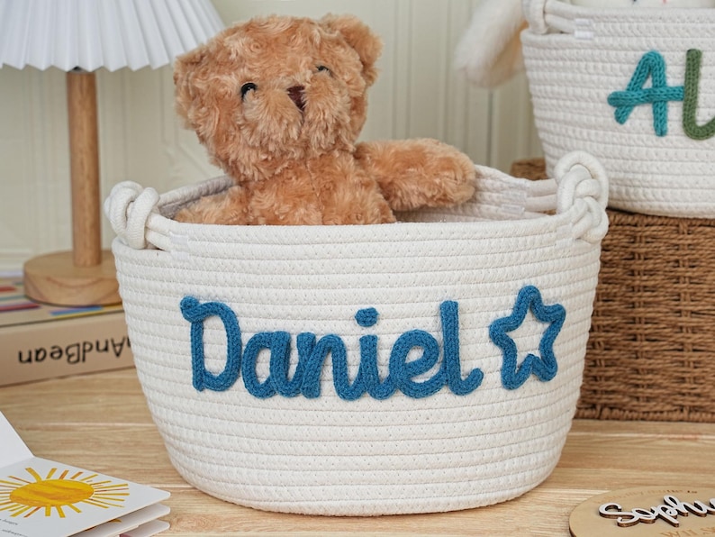 May include: A white rope basket with the name "Daniel" in blue script and a star. A brown teddy bear sits inside the basket. Another basket with the name "Ali" is visible in the background. The baskets are for storage.