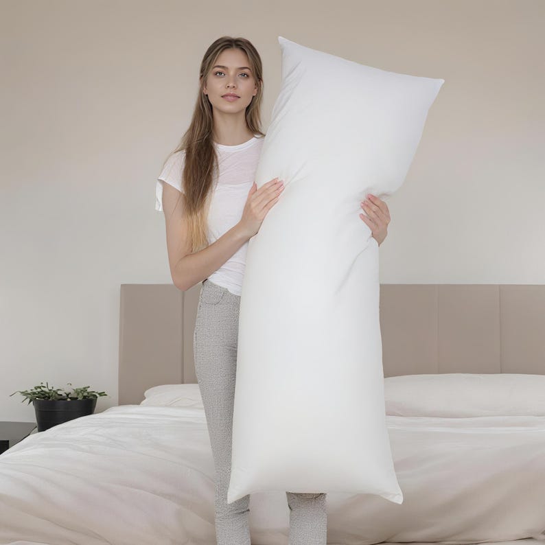 May include: A long, white body pillow held by a person. The pillow is rectangular and appears to be filled with a soft material. The person is standing in front of a bed with white bedding. The pillow is approximately 152 cm long.