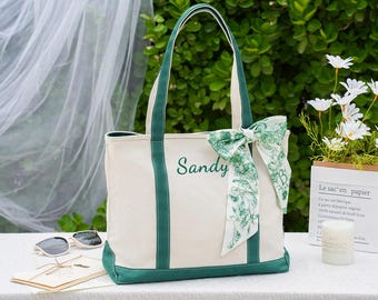 May include: Cream-colored tote bag with green accents, handles, and base. The word "Sandy" is embroidered in green. A decorative scarf with a green floral pattern is tied to the handle. Sunglasses and a card are on the table.