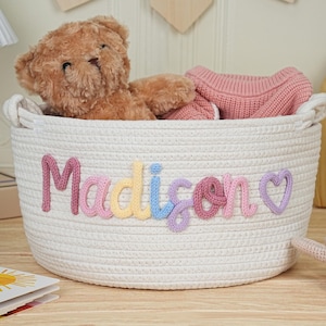 May include: A white woven storage basket with the name "Madison" spelled out in colorful yarn letters. The basket contains a teddy bear and a pink sweater. A wooden picture frame and a book are in the background.