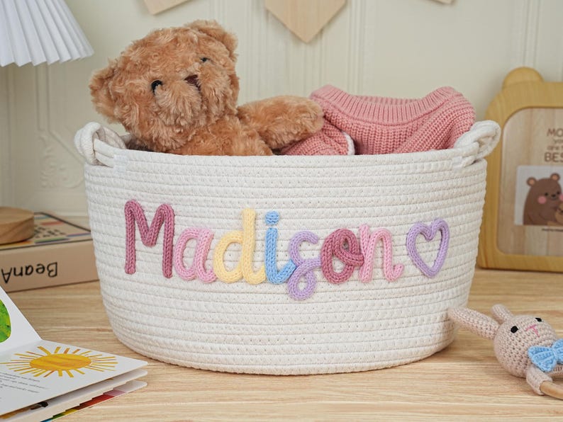 May include: A white woven storage basket with the name "Madison" spelled out in colorful yarn letters. The basket contains a teddy bear and a pink sweater. A wooden picture frame and a book are in the background.