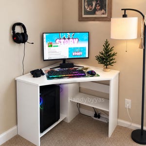 May include: A white corner computer desk featuring a black desktop computer tower, a monitor displaying the Pet Story game interface, a backlit keyboard, a gaming mouse, and black headphones mounted on the wall. A small artificial pine tree sits nearby.