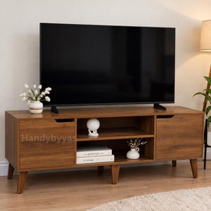 May include: A rectangular wooden media console with a medium brown wood grain finish, featuring two side cabinets with cutout handles, two open central shelves, and five tapered legs. A large black television sits on top of the console.