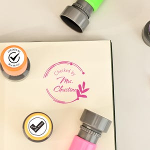 May include: A selection of self-inking stamps in shades of pink, orange, and green. The stamps are positioned around a cream-coloured surface with the text "Checked by Mrs. Christine" and other text. These stamps are designed for marking documents.
