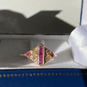 Puede incluir: Un anillo de oro con un diseño geométrico. El anillo presenta una fila central de piedras rojas cuadradas flanqueadas por gemas rojas más pequeñas y detalles de diamantes. El anillo está en una caja de joyería azul.