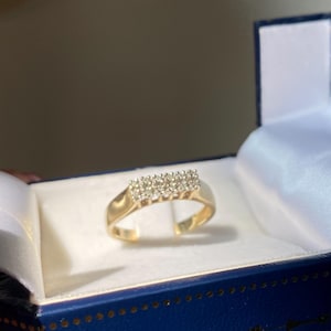 Peut inclure: Une bague en or avec un groupe rectangulaire de petites pierres claires. La bague est présentée dans un écrin bleu avec un intérieur en satin blanc. La boîte a une bordure dorée.