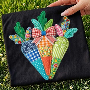 May include: A black fabric square featuring a colorful applique of three carrots. Each carrot is made of patchwork fabric in various patterns and colors, including blue gingham, orange polka dots, and green stripes. A red and white checkered bow sits atop the carrots.