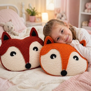 Op de afbeelding: Twee gehaakte vossen-vormige kussens. De ene is dieprood, de andere oranje, beide met crèmekleurige gezichten, zwarte ogen en neuzen. De kussens liggen op een bed met een roze dekbed. De kussens zijn ongeveer 36 cm breed.