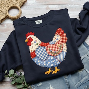 May include: Navy blue sweatshirt featuring a patchwork chicken design. The chicken has a red comb and wattle, a yellow beak, and a body made of various patterned fabrics, including gingham and floral prints. The sweatshirt has a crew neck and long sleeves.