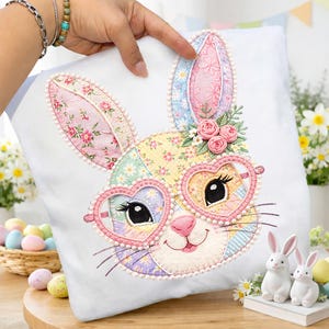 May include: A white square pillow featuring an embroidered bunny face with heart-shaped glasses. The bunny's face is a patchwork of floral fabrics in pastel colors. The design includes pink roses and pearl-like trim, perfect for Easter decor.