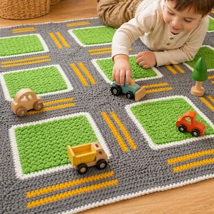 May include: A crocheted play mat designed as a road with green squares and yellow road markings. Wooden toy cars and a tree are placed on the mat. The mat is gray, green, white, and yellow. A child is playing with the toys on the mat.