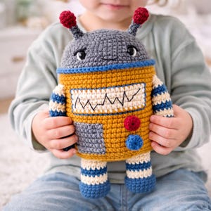 Puede incluir: Un juguete de robot de ganchillo hecho a mano en tonos amarillo, gris, azul y rojo. El robot tiene un cuerpo rectangular con un diseño cosido, brazos y piernas a rayas, y elementos tipo antena en la cabeza. El juguete es sostenido por un niño.