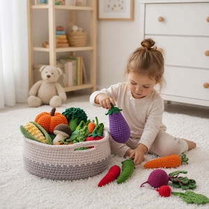 Puede incluir: Una niña juega con una cesta de verduras de ganchillo, que incluye una calabaza, brócoli, maíz, zanahorias y una seta. La cesta es gris y blanca, y las verduras son de colores brillantes. La niña sostiene una berenjena morada.