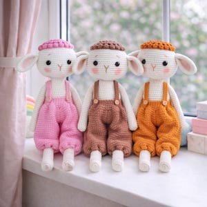 May include: Three handmade crochet sheep dolls with white bodies, pink, brown, and orange overalls, and matching hats. Each doll has a smiling face with rosy cheeks and button details on the overalls. They are sitting on a windowsill.