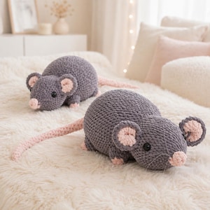 May include: Two gray crochet rat plush toys with pink ears, noses, and tails. The stuffed animals are lying on a white, fluffy surface. The rats have black eyes and are made of a soft, textured yarn.