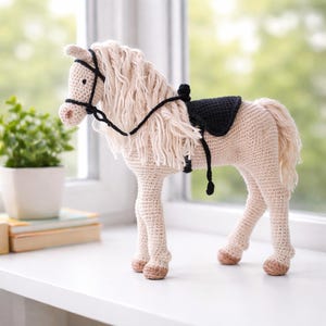Puede incluir: Figura de caballo de ganchillo en crema y beige. El caballo tiene una larga melena y cola sueltas, una brida negra y una silla de montar negra. Está sobre una superficie blanca cerca de una ventana, con una pequeña planta en maceta al fondo.