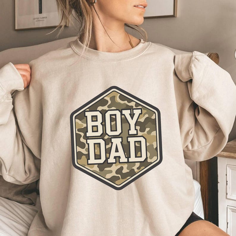 a woman sitting on a bed wearing a sweatshirt that says boy dad