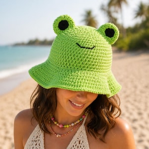 May include: A vibrant green crochet bucket hat, shaped like a frog, with black eyes and a smiling mouth. The hat is worn by a person on a sandy beach, with a sea view.