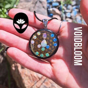 Puede incluir: Un collar con un colgante negro con un diseño dorado de flor de la vida y piedras de ópalo iridiscentes. El colgante está en un cordón negro.