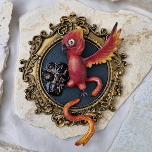 Könnte beinhalten: Eine rote und gelbe Phönix-Figur mit ausgebreiteten Flügeln, die auf einem schwarzen runden Sockel mit einem goldenen verzierten Rahmen sitzt. Der Phönix ist mit einem feurigen Schwanz und einem schwarzen und roten Pfotenabdruck dargestellt.
