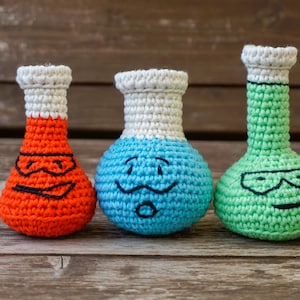 May include: Three crocheted beaker-shaped toys with different colored faces. The beakers are orange, blue, and green. The orange beaker has a black eye mask, the blue beaker has a mustache and a surprised expression, and the green beaker has a black eye mask and a smiling expression.