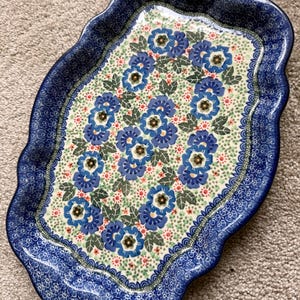 May include: An oval-shaped ceramic serving platter with a scalloped edge. The platter features a blue floral pattern on a cream background, with a dark blue border. The floral design includes blue flowers, green leaves, and red accents.