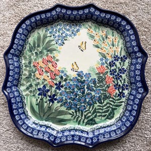 May include: A decorative ceramic serving platter with a scalloped edge. The platter features a floral design with butterflies in the center, surrounded by a border of blue and white floral patterns. The colors include blue, green, yellow, and coral.