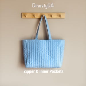 May include: A light blue quilted tote bag with vertical stitching, hanging from a wooden rack. The bag has two handles and the text "Zipper & Inner Pockets" is visible. The brand name "DinastyUA" is at the top.