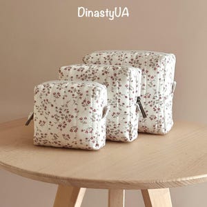 May include: Three quilted cosmetic bags in different sizes, patterned with a white background and a red floral design. Each bag has a zip closure and small side handles. The bags are displayed on a light-coloured wooden table. The text "DinastyUA" is visible.