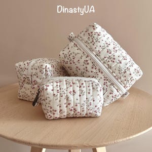 Op de afbeelding: Drie gewatteerde make-up tassen met een bloemenpatroon in rood en wit, gerangschikt op een houten tafel. De tassen zijn verschillend van grootte, met ritsen en een gewatteerde textuur. De tekst "DinastyUA" is zichtbaar.