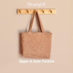 May include: A tan corduroy tote bag with long handles hangs from a wooden rack. The bag has a rectangular shape and is made of a ribbed fabric. The text "Zipper & Inner Pockets" is visible at the bottom of the image. The brand name "DinastyUA" is at the top.