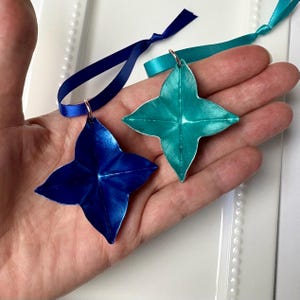 May include: Two star-shaped ornaments, one blue and one teal, with metallic finishes. Each ornament has a small loop and is attached to a satin ribbon. The ornaments are held in a hand, with a white background.