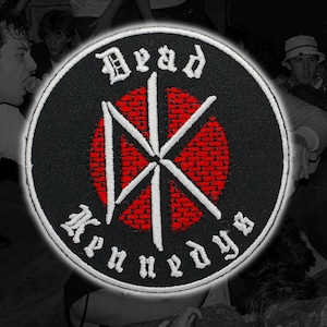 May include: A circular embroidered patch featuring a black background with a red brick wall pattern in the centre. A white stylised DK logo is overlaid on the red circle. The text Dead Kennedys is embroidered in white gothic font along the border.
