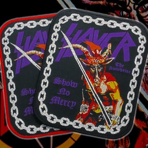 May include: Two black patches with a silver chain border. The patches feature the word "SLAYER" in purple, a horned figure, and the text "Show No Mercy" and "The Antichrist". One patch has a red border.
