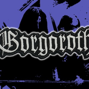 May include: Embroidered patch with the word "Gorgoroth" in white gothic lettering on a black background. The patch has a black border and is set against a purple and black abstract design.