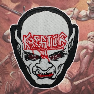 May include: Embroidered patch with a demonic face and the word "KREATOR" in red. The patch has a black border and is set against a background of a battle scene with skeletons and figures.