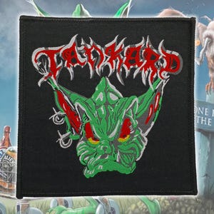 May include: Black square patch with the word "TANKARD" in red, stylized lettering. A green, monster-like creature with red accents is centered below the text. The patch is likely for a jacket or bag.