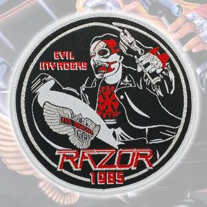 Puede incluir: Un parche bordado circular con fondo negro y borde blanco. Representa una figura con chaqueta de cuero sosteniendo un arma, con el texto "EVIL INVADERS" y "RAZOR 1985" en rojo. Un emblema alado es visible en la parte inferior izquierda.