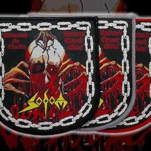 Peut inclure: Trois écussons brodés sur fond noir, représentant un crâne et les textes "Obsessed By Cruelty" et "Teutonic Thrash Metal". Les écussons ont une bordure en chaîne et sont en noir, gris et rouge.