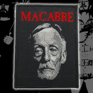 May include: A rectangular black fabric patch featuring an embroidered portrait of a man in shades of gray and white. The word MACABRE is embroidered in bold red capital letters across the top of the patch. The edges are finished with black stitching.