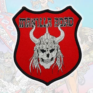 Puede incluir: Un parche bordado en forma de escudo con fondo rojo y borde negro. El parche presenta una calavera plateada con un casco con cuernos y el texto MANILLA ROAD en una fuente estilizada en blanco y negro en la parte superior central.