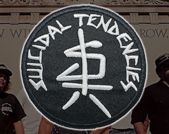 May include: A circular black embroidered patch featuring white stitching. The text "SUICIDAL TENDENCIES" is arched along the top and bottom border. In the center, a white stylized graphic symbol resembles a combination of the letters S and T.