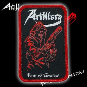 以下が含まれることがあります： 赤い縁取りのある黒い長方形のパッチ。「Artillery」の文字が白い筆記体で書かれています。武器を持った赤いフード付きの人物が下に刺繍されています。「Fear of Tomorrow」の文字が下部にあります。