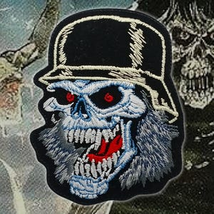 Könnte beinhalten: Gestickter Aufnäher mit einem Totenkopf, der einen schwarzen Hut trägt. Der Schädel ist in Blau und Weiß mit roten Augen und einem roten Mund detailliert. Der Aufnäher hat einen schwarzen Rand, für Kleidung oder Accessoires.